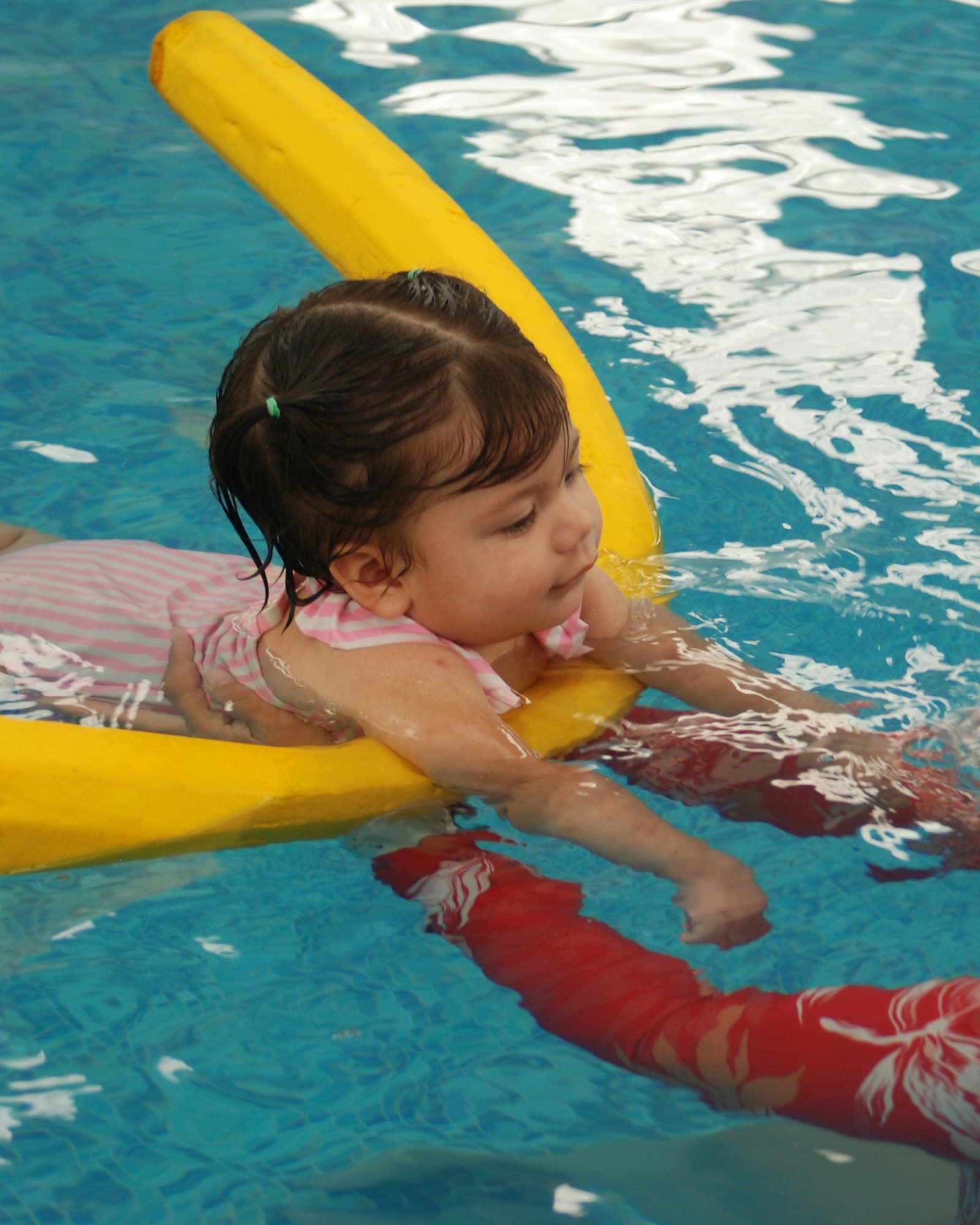 A young child resting on a pool noodle in bright blue water, steadied by an instructor's guiding arm.