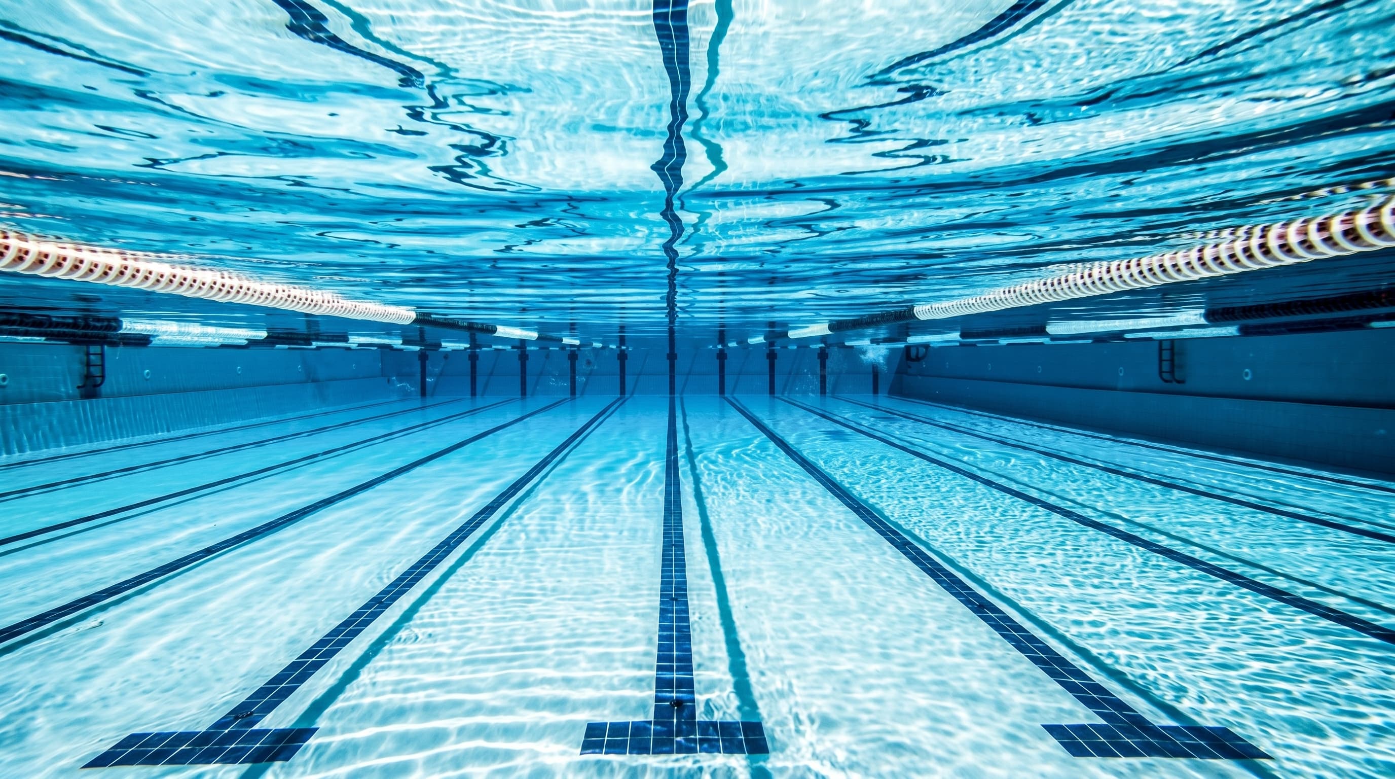 Underwater view of a competition pool, lane lines stretching into the distance beneath shimmering blue water.